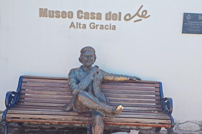 A bronze statue of a seated man on a bench with a plaque reading 'Museo Casa del Le Alta Gracia' in the background. 2
