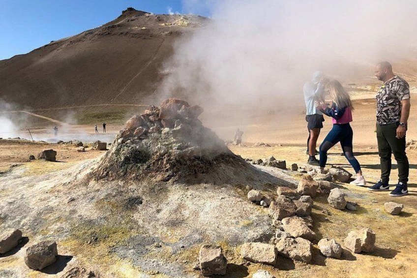 Un área geotérmica con vapor ascendiendo desde el suelo, terreno rocoso y una gran colina al fondo. 3