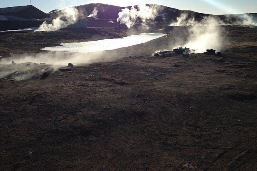 Un área geotérmica con fumarolas, un lago pequeño y montañas al fondo. 4
