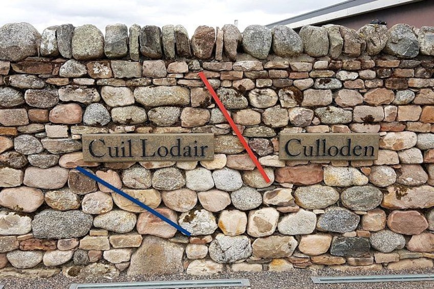 A stone wall with two signs, one labeled 'Cuil Lodair' and the other 'Culloden'. 2