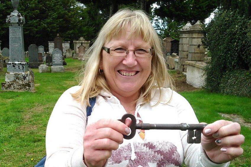 A person holding a key in a cemetery with gravestones and a tree in the background. 2
