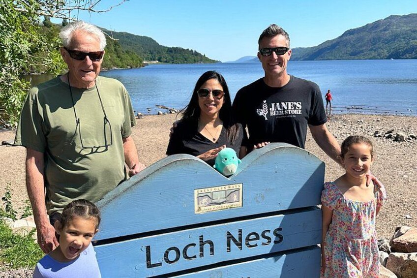 A family standing by a sign that reads 'Loch Ness' with a lake and mountains in the background. 2