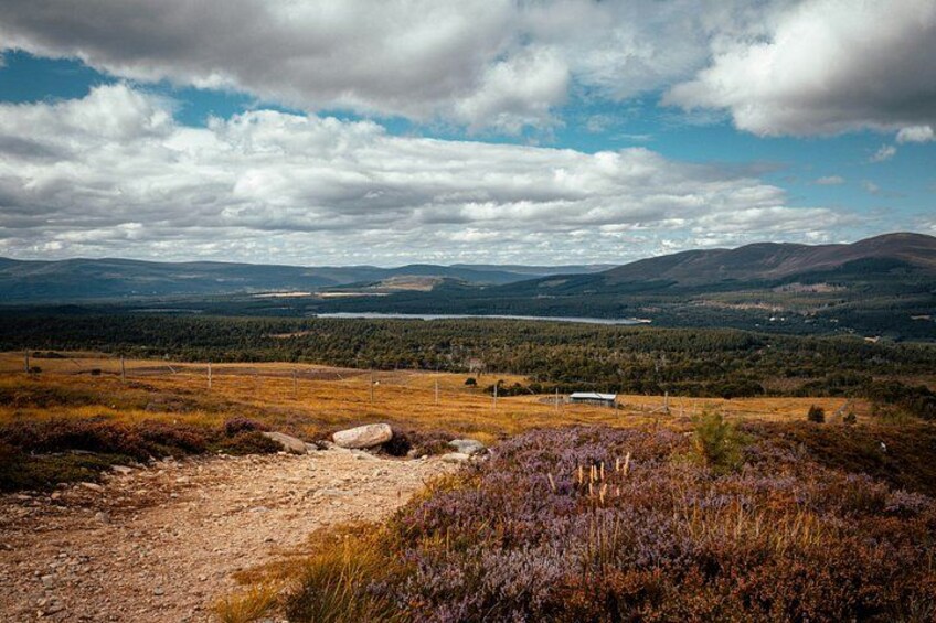 Een bergachtig landschap met een onverhard pad, schaarse begroeiing en een meer in de verte. 1