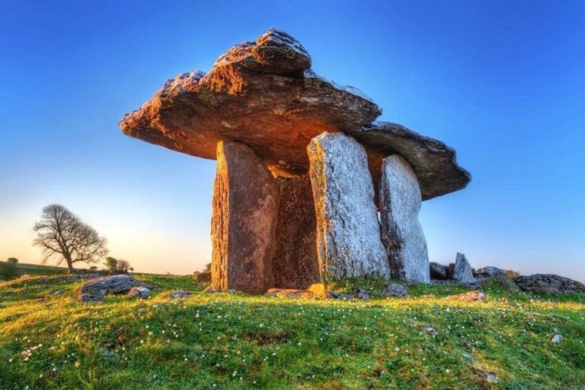 A stone circle with large upright stones and a smaller stone at the top, set in a grassy field with a clear sky. 3