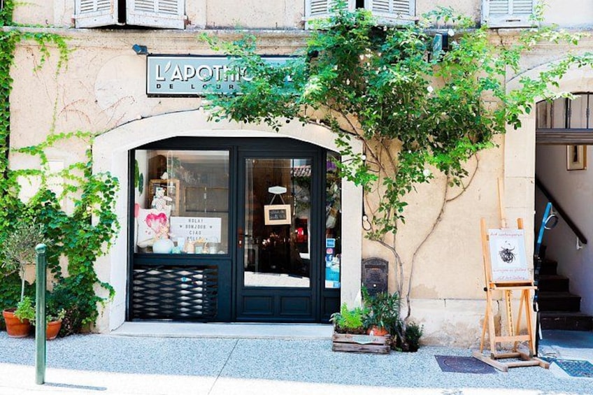 A storefront with a sign reading 'L'APOTHICIE DE LOURT,' a window display, and a small easel with a painting. 5