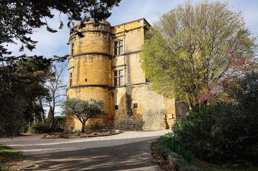 A historic stone building with a cylindrical tower, surrounded by trees and a paved driveway. 3