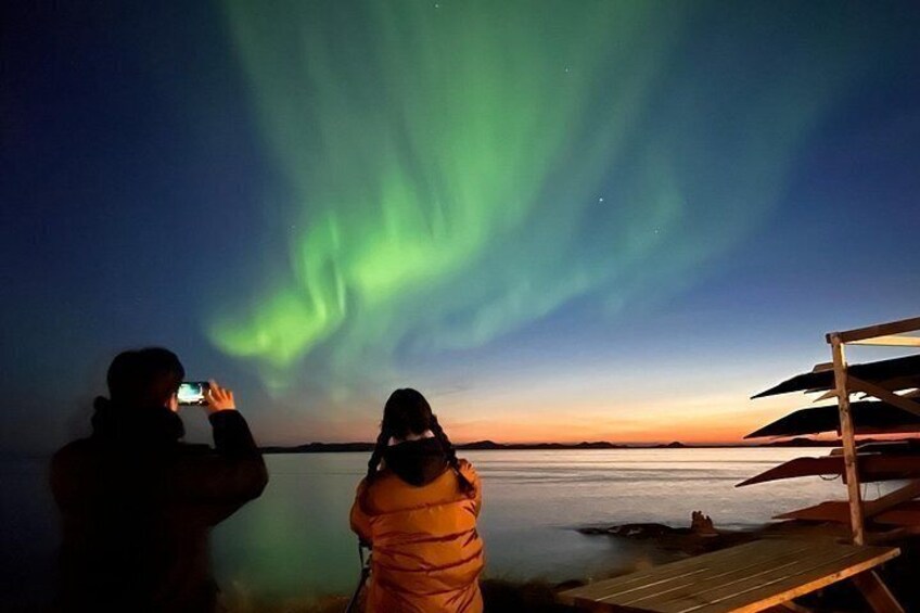 To personer fotograferer nordlys over vandet i skumringen. 3