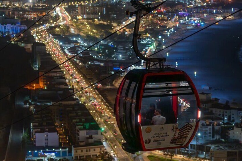A cable car with passengers, a brightly lit cityscape below, and a coastline in the distance. 3