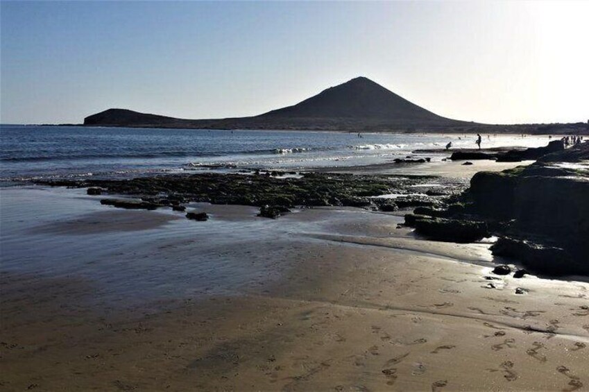 Ein Strand mit Fußabdrücken, eine felsige Küstenlinie und ein Berg im Hintergrund. 1