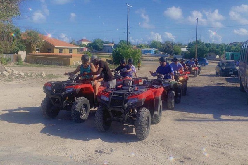 A group of people on ATVs on a dirt road with buildings in the background. 4