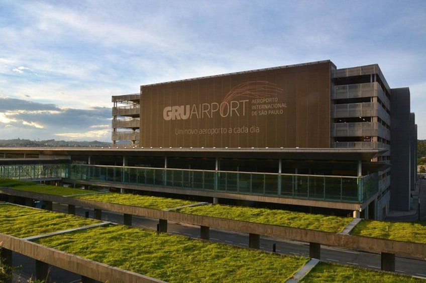 A modern airport terminal with a large sign reading 'GRU AIRPORT' and a green rooftop garden. 3