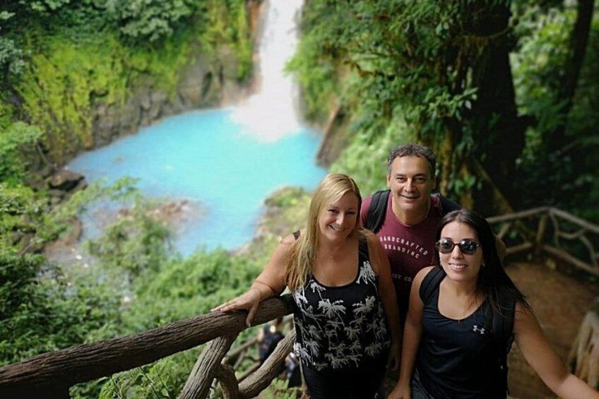 Three people on a wooden bridge with a river and greenery in the background. 1