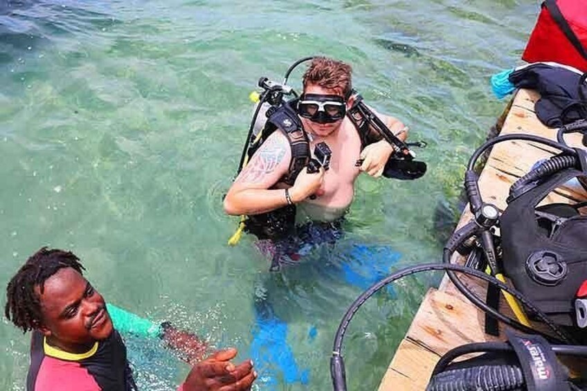 Une personne vêtue d’une combinaison de plongée et portant un réservoir d’air, une autre personne portant un t-shirt rose, et un bateau en bois équipé de matériel de plongée. 3