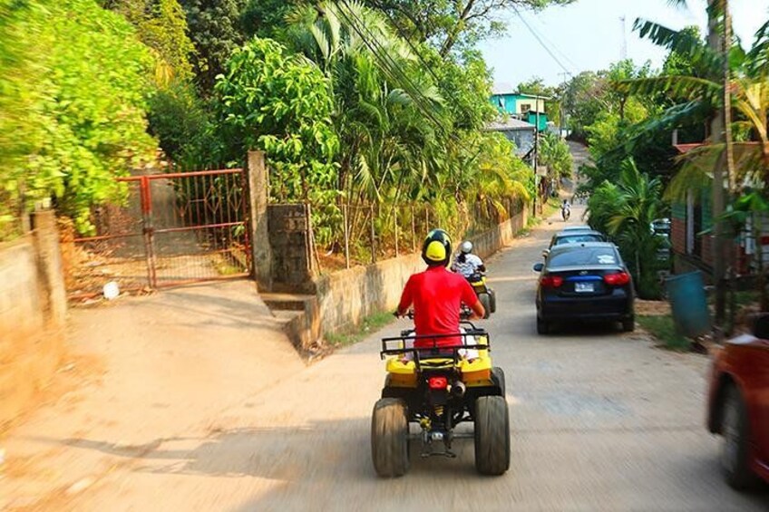 A person riding a quad bike on a narrow road with cars parked on the side, surrounded by greenery and a metal gate. 4