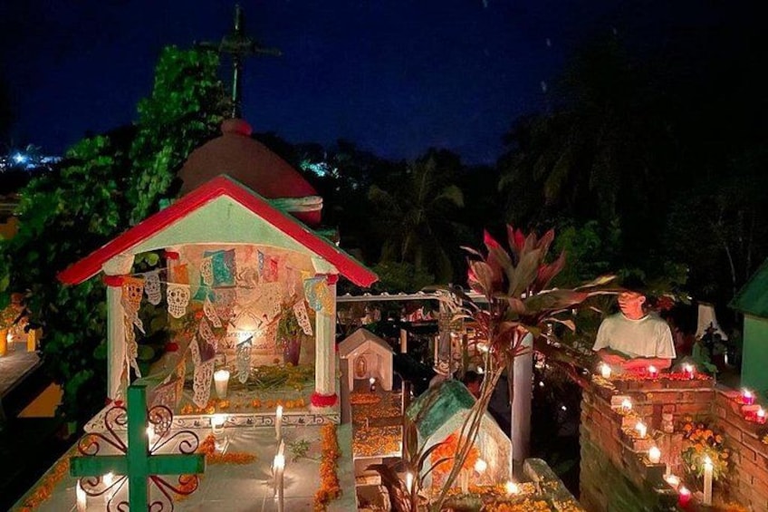 Santuario religioso nocturno al aire libre con una pequeña capilla, velas y una estatua. 4