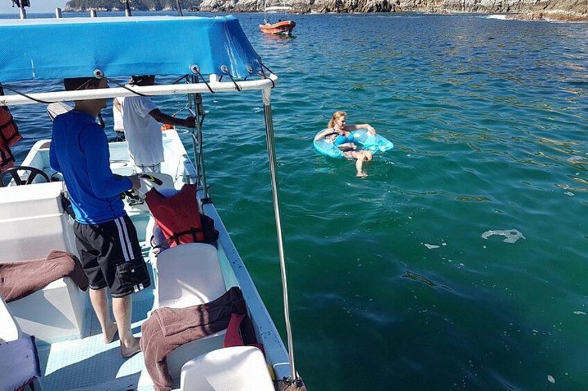 Un bote con una persona con camisa azul y otra con camisa marrón, un niño sobre un flotador azul, y agua tranquila. 4