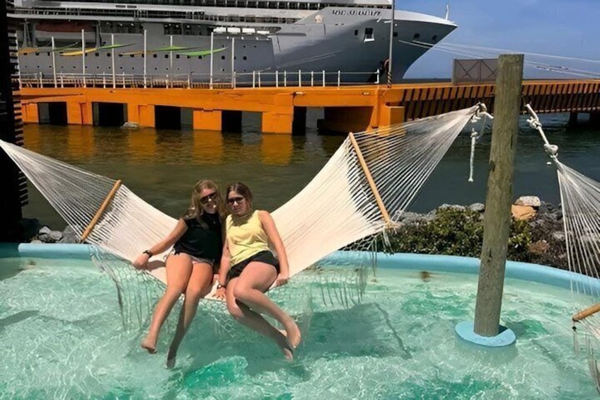Two people sitting on a hammock in a pool with a cruise ship in the background. 2