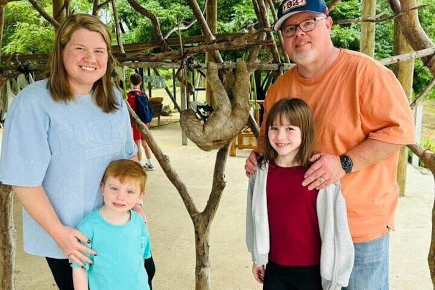 A family of three standing under a wooden structure with a sloth hanging from a branch. 4