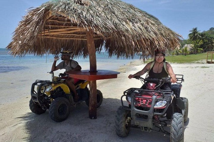 Deux personnes sur des quads, sur une plage, à l’ombre d’un parasol en chaume. 2