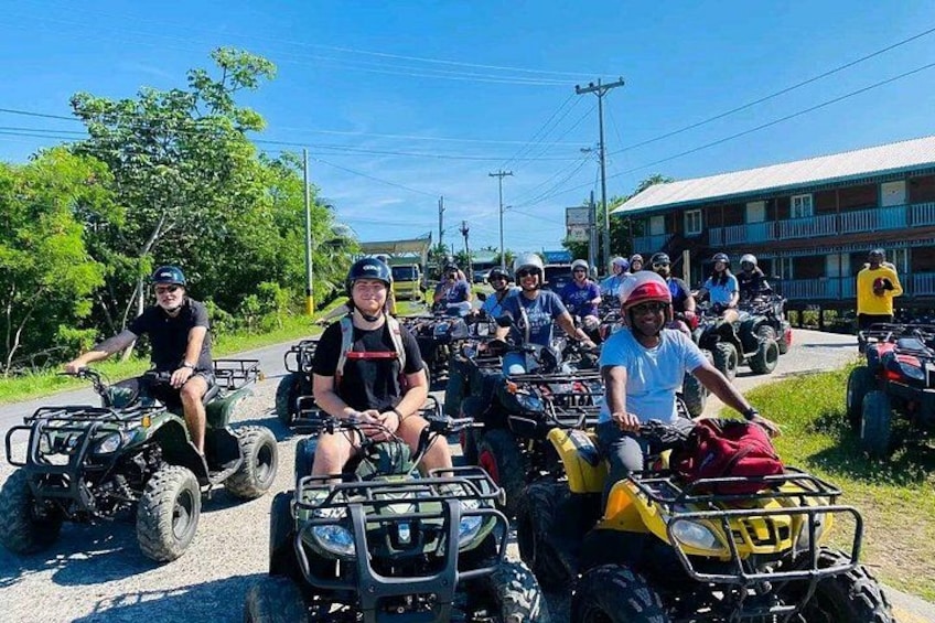 Un groupe de personnes circulant en quads sur une route, avec en arrière-plan de la végétation et des bâtiments. 1
