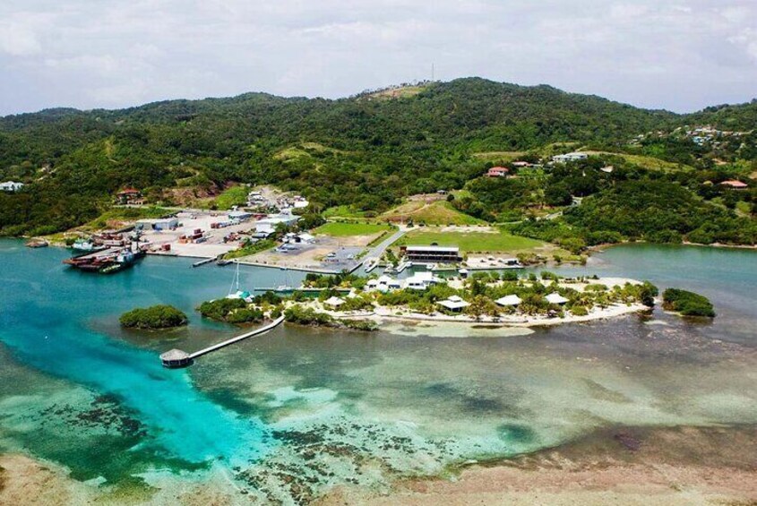 An aerial view of a coastal resort with clear turquoise waters, a dock, and lush green hills in the background. 2