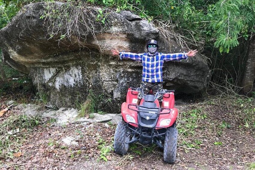 A person on a red ATV with arms outstretched in a forested area with large rocks and greenery. 1