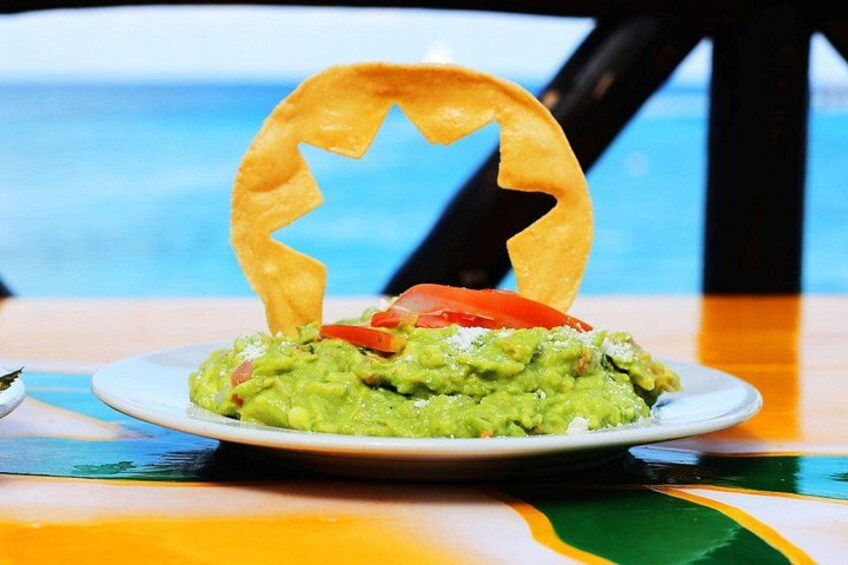A plate of guacamole with a tortilla chip shaped like a star, a slice of tomato, and a small bowl of salsa on a table with a colorful tablecloth. 4