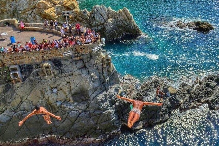 A rocky coastline with people sitting on a ledge and two individuals diving into the water. 1