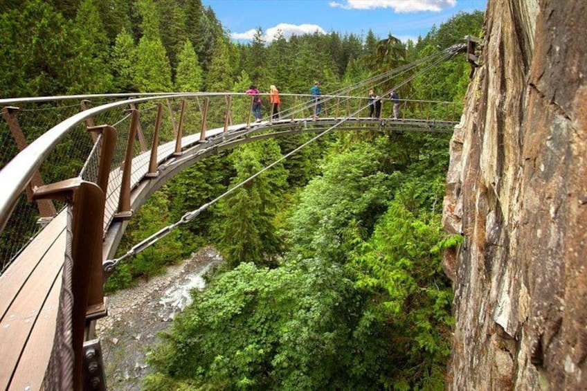 Un puente colgante con gente caminando sobre él, rodeado de un bosque frondoso y un acantilado rocoso. 5