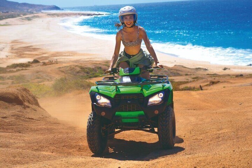 Une personne est assise sur un quad vert sur un chemin de terre près d’une plage, avec l’océan en arrière-plan. 3