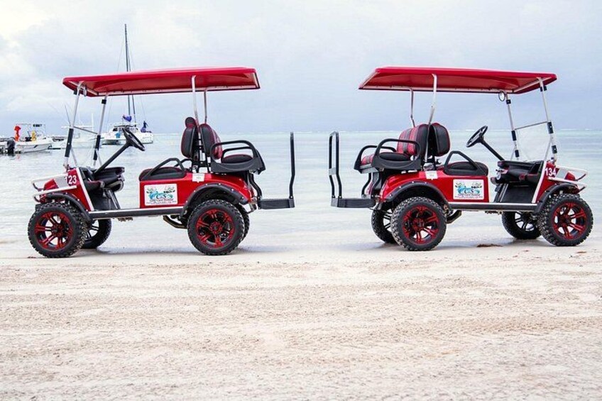 To røde golfbiler med sorte sæder og hjul på en sandstrand med havet i baggrunden. 3