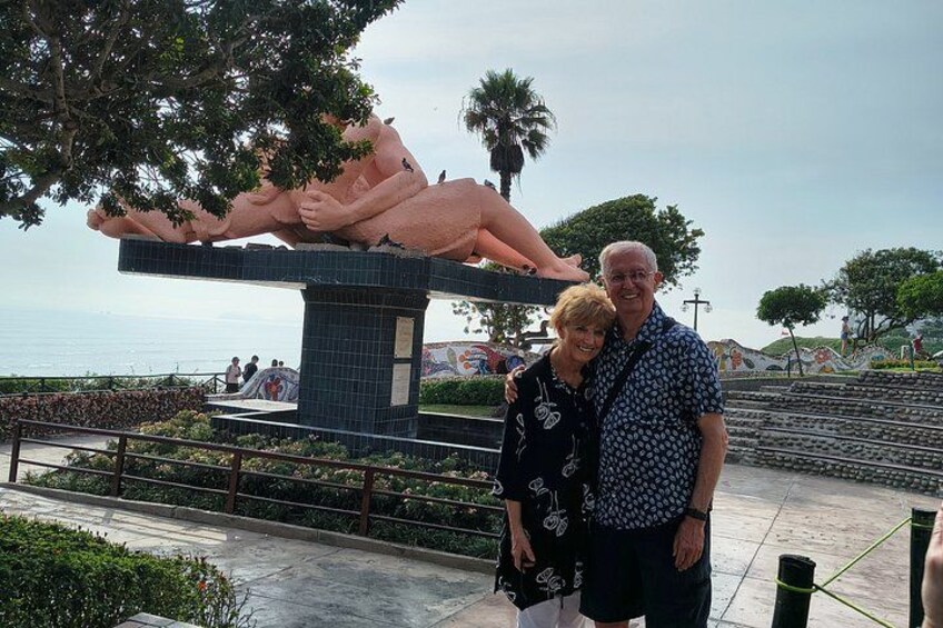 Una pareja posa frente a una gran escultura, con un árbol y una masa de agua al fondo. 1