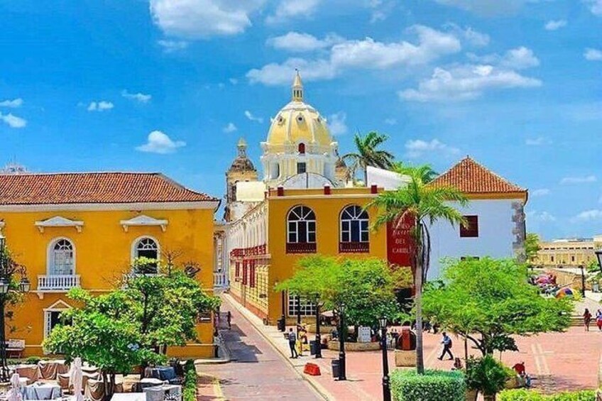 A historic building with a yellow facade and a dome, surrounded by greenery and palm trees. 1