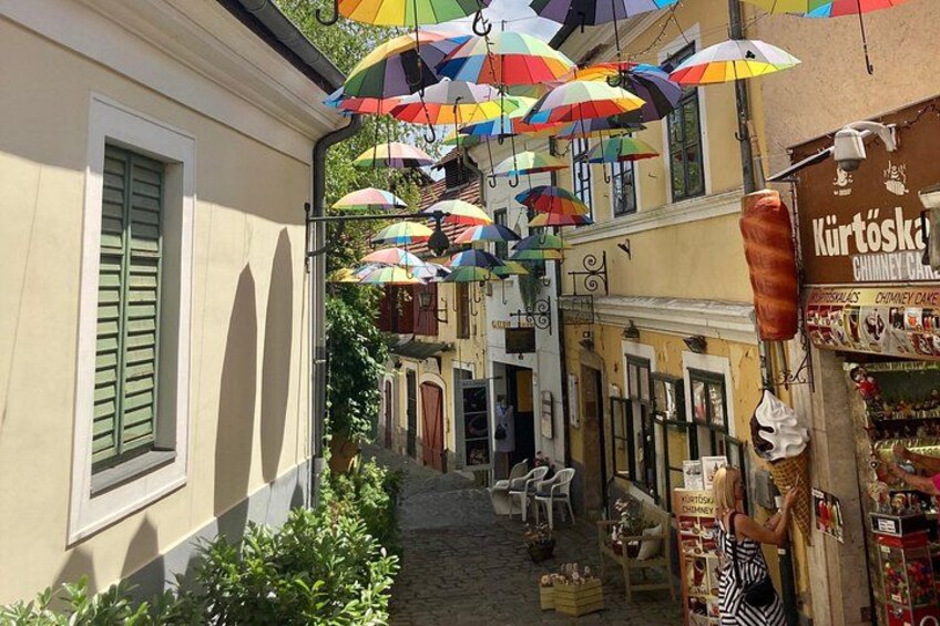 Una calle estrecha con paraguas de colores colgando, una tienda de kürtőskalács y un edificio con contraventanas verdes. 4