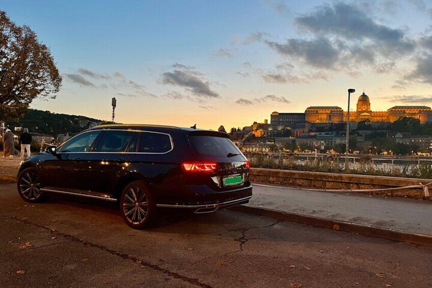 Un coche negro estacionado al borde de una carretera, con un paisaje urbano y un edificio grande al fondo. 1