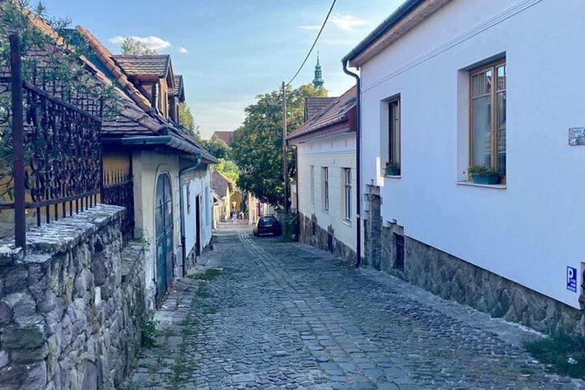 Eine gepflasterte Straße gesäumt von traditionellen Gebäuden, ein geparktes Auto und ein Kirchturm in der Ferne. 2