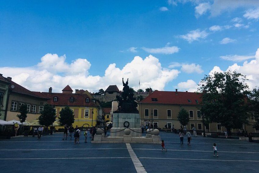 A public square with a statue, buildings, and people. 3