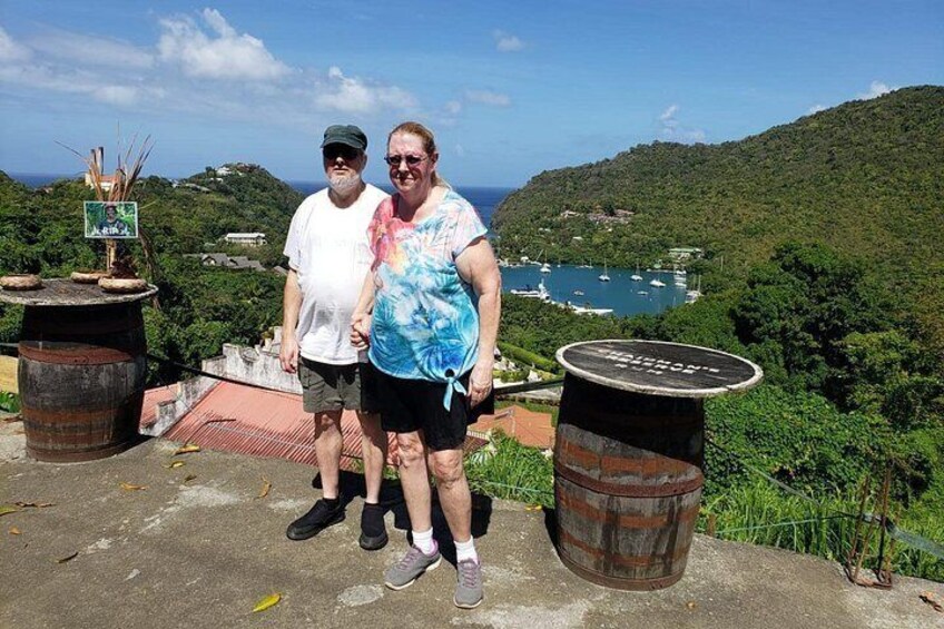 Two people standing on a concrete platform with a scenic view of a coastal area, including a hill, trees, and a body of water with boats. 3