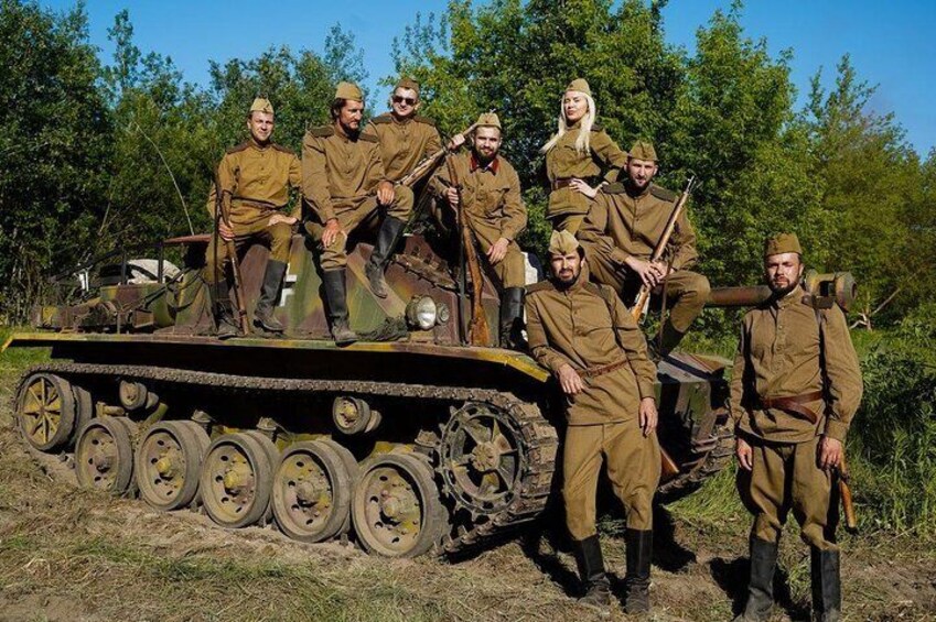 A group of people in military uniforms posing on a tank in a wooded area. 4