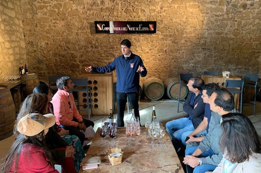 A group of people seated around a table with wine bottles and glasses, in a rustic room with stone walls and barrels. 3