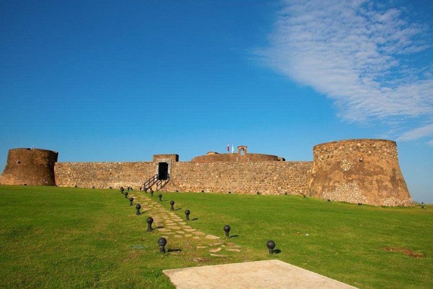 A historic fort with a grassy pathway leading to the entrance, surrounded by stone walls. 2