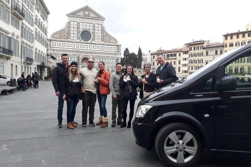 Un gruppo di persone in piedi di fronte a un edificio con una cupola e colonne, una macchina nera parcheggiata nelle vicinanze e un panorama urbano sullo sfondo. 5