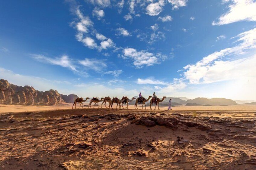 A caravan of camels in a desert landscape with rocky terrain and distant mountains. 4