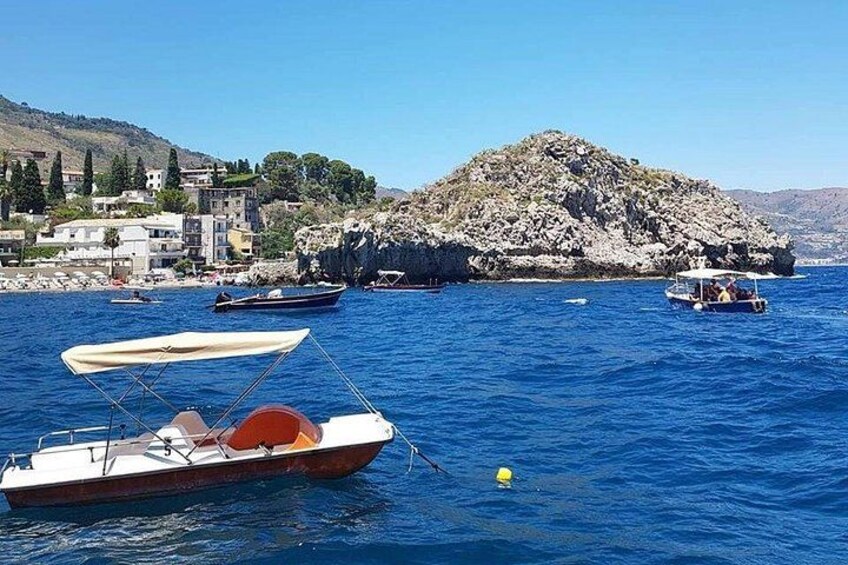 A coastal scene with boats on calm blue waters, rocky outcrops, and buildings on the shore. 3