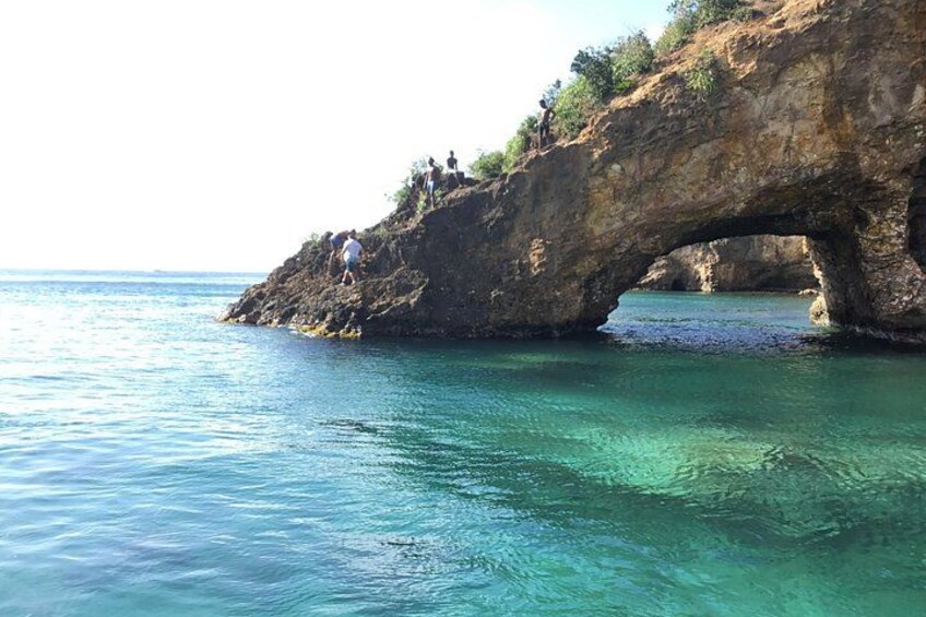 A natural rock arch formation over clear blue water with people climbing on the rock. 1