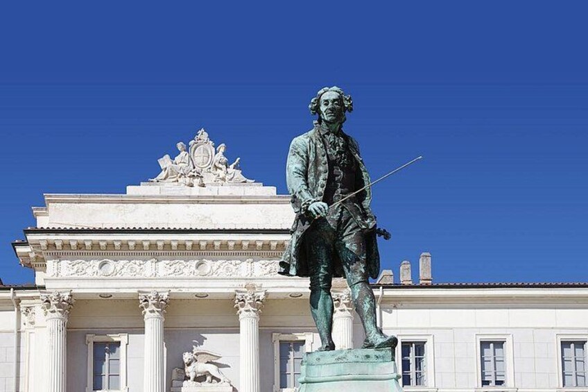 A statue of a historical figure in period clothing, standing on a pedestal in front of a classical building with columns and decorative elements. 4