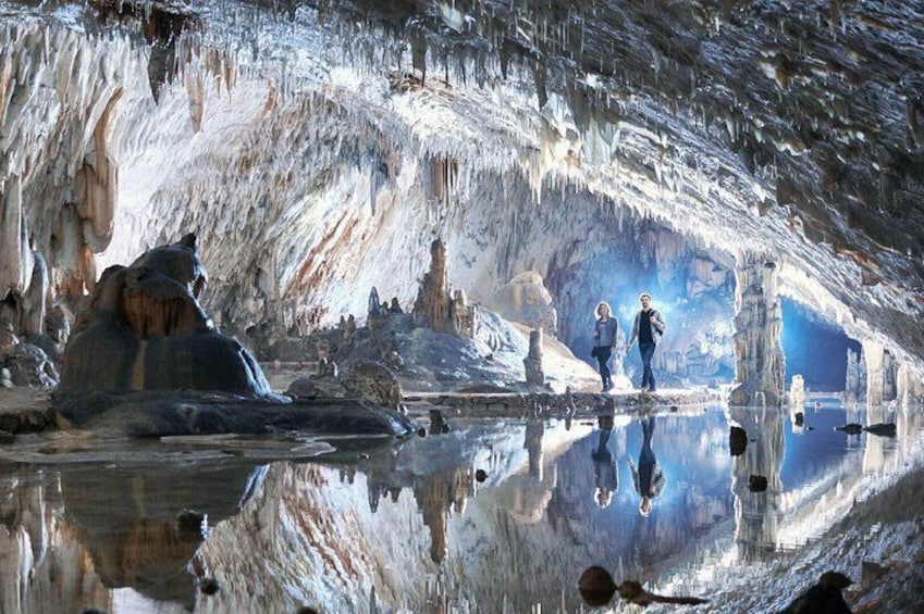 An ice cave with stalactites and stalagmites, a reflective pool, and two people standing in the center. 4