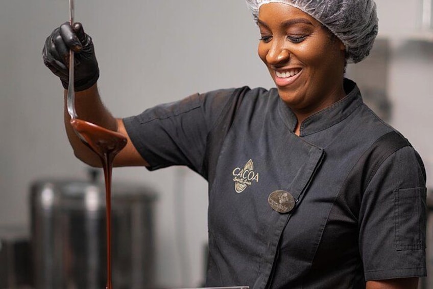 A person wearing a black chef's uniform with a logo, holding a ladle with a thick, dark liquid. 1