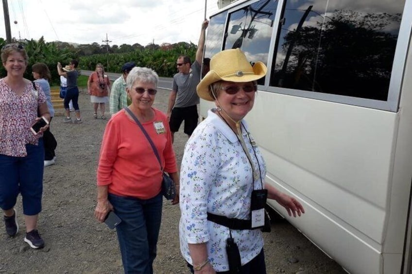 People standing near a bus, some wearing hats, in a rural setting with greenery in the background. 5