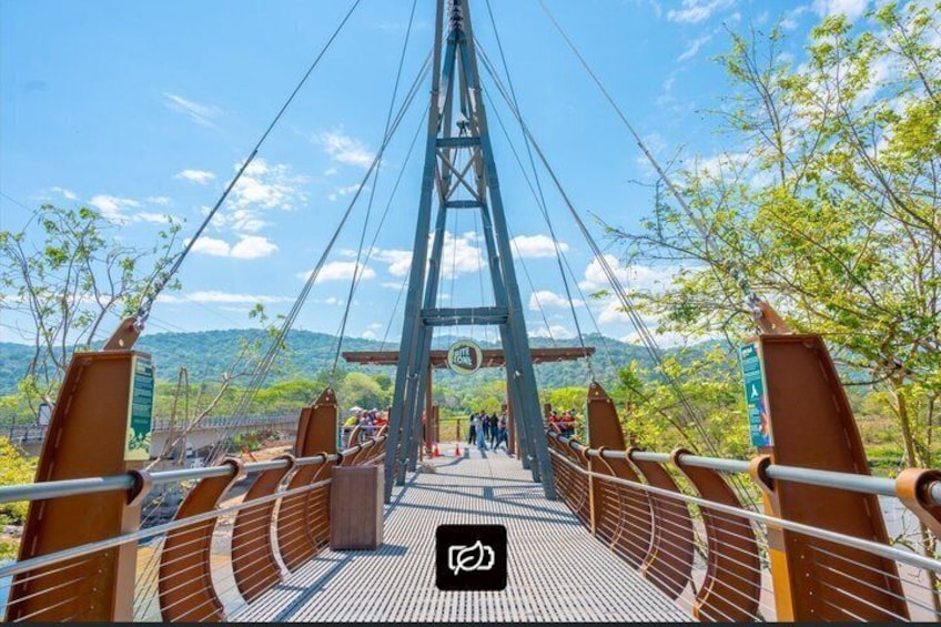 A suspension bridge with a metal framework and wooden planks, surrounded by greenery and mountains in the background. 3